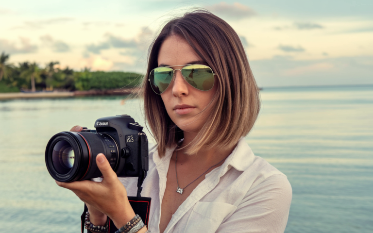 A photo of a woman with shoulder-length brown hair wearing aviator sunglasses and a white shirt. She holds a Canon DSLR camera with a lens attached, positioned close to her face. The background reveals a serene tropical beach setting with calm waters and a clear sky. The woman's accessories include a necklace and multiple bracelets.