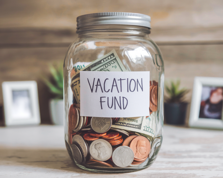 Vacation Fund Jar sitting on desk with coins and cash in it. Saving for travel plans.