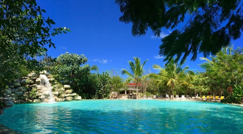 This image shows a beautiful tropical resort swimming pool in Punta Cana, Dominican Republic. The large turquoise pool features a rock waterfall on the left side. Surrounding the pool are lush palm trees and tropical vegetation under a bright blue sky with a few white clouds. In the background, there are lounge chairs along the pool deck and what appears to be a small resort building with a red roof. The scene captures the idyllic vacation atmosphere typical of Caribbean resorts with its clear water, landscaped grounds, and pristine conditions. One of Punta Cana's hidden gems to go visit.
